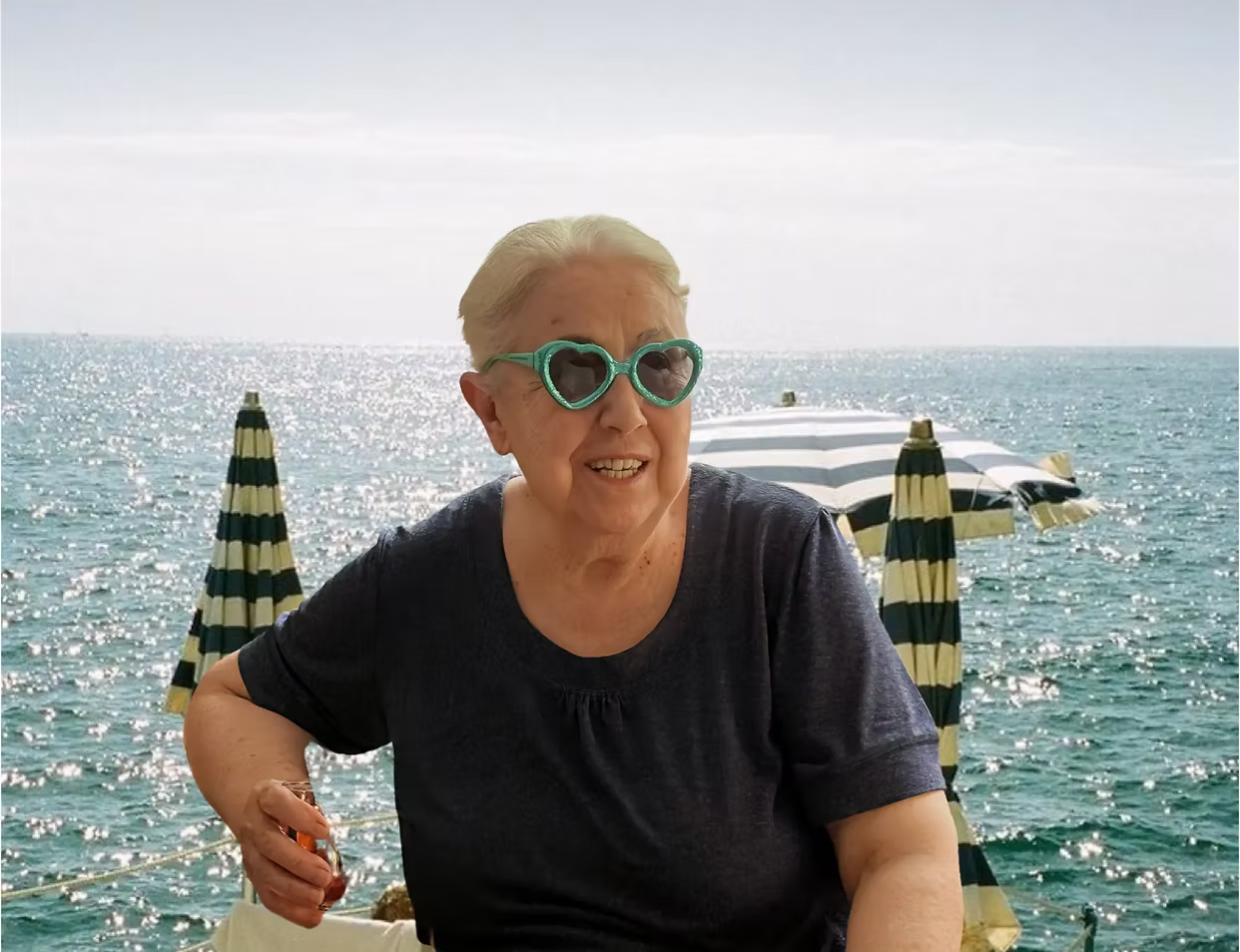 Person wearing heart-shaped sunglasses sitting by the ocean with striped umbrellas.