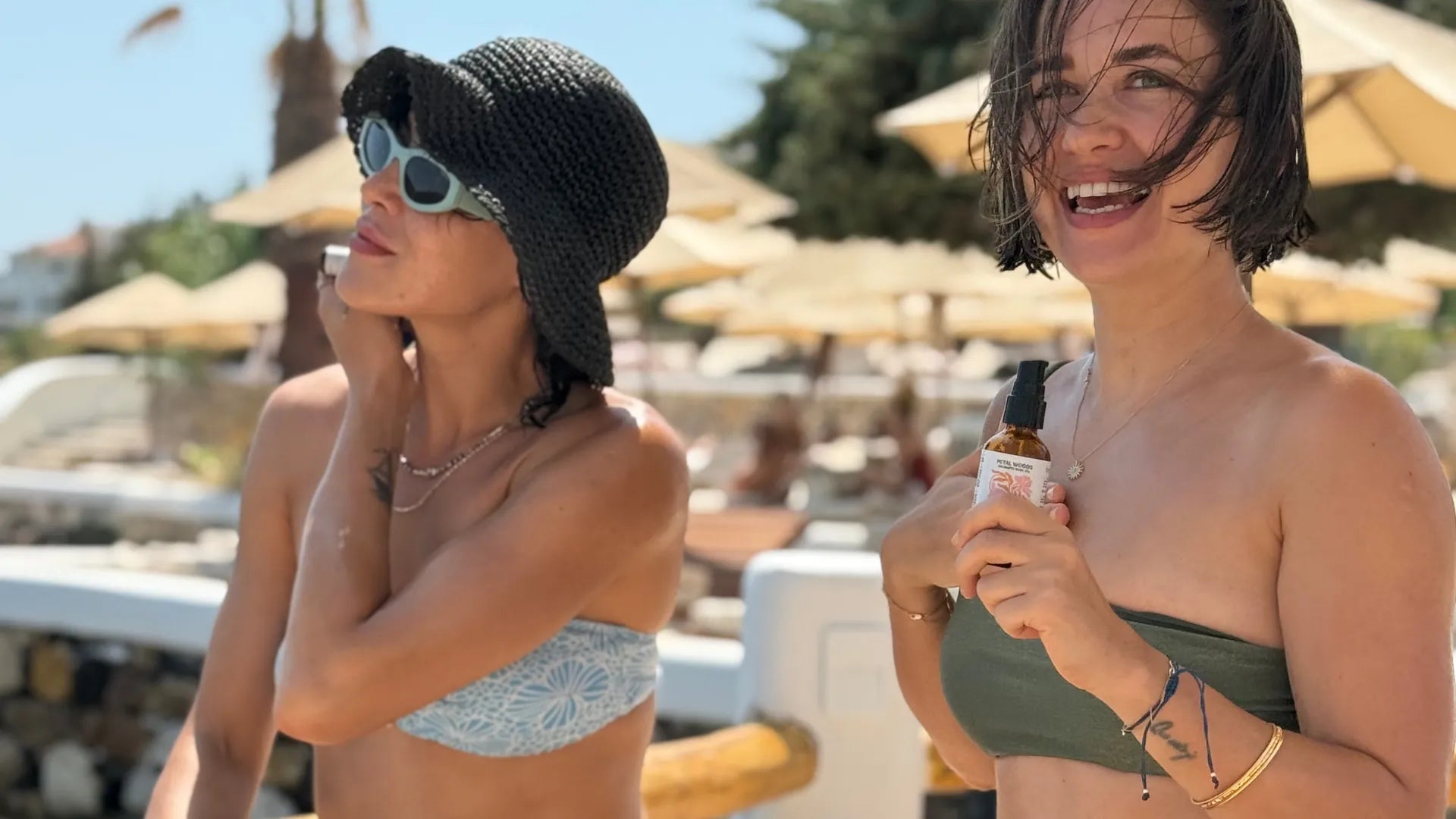 Two women on a beach, one applying sunscreen and the other holding a bottle.